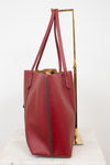 A red leather tote bag with a pebbled texture, displayed on a white marble surface next to a gold metal stand against a white background.