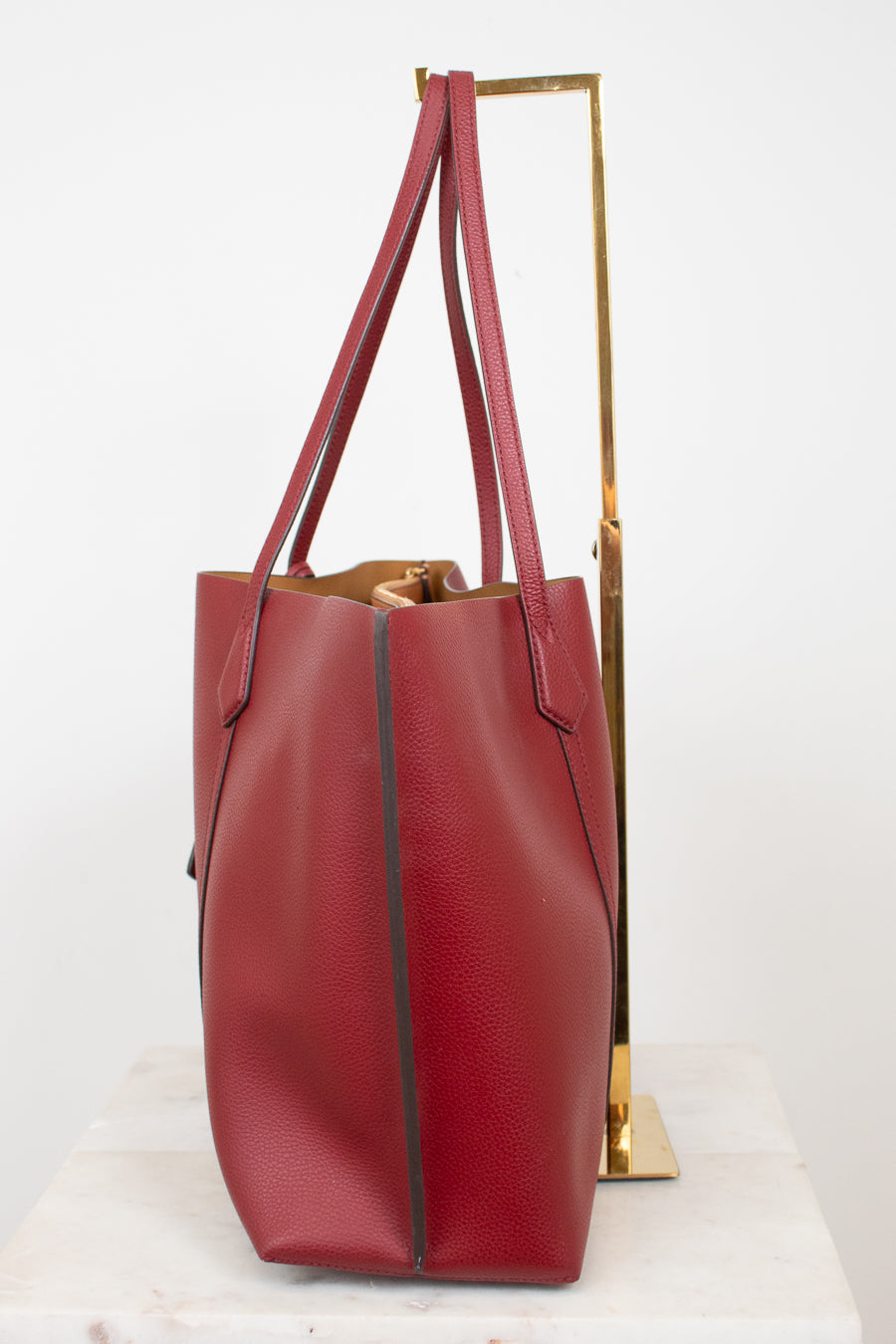 A red leather tote bag with a pebbled texture, displayed on a white marble surface next to a gold metal stand against a white background.
