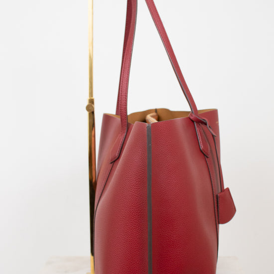 A red pebbled leather tote bag with double shoulder straps, displayed on a gold metal stand against a white background.