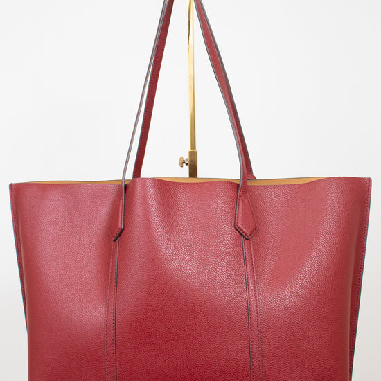A red pebbled leather tote bag with dark red handles and gold-tone hardware, displayed on a white marble block against a white background.