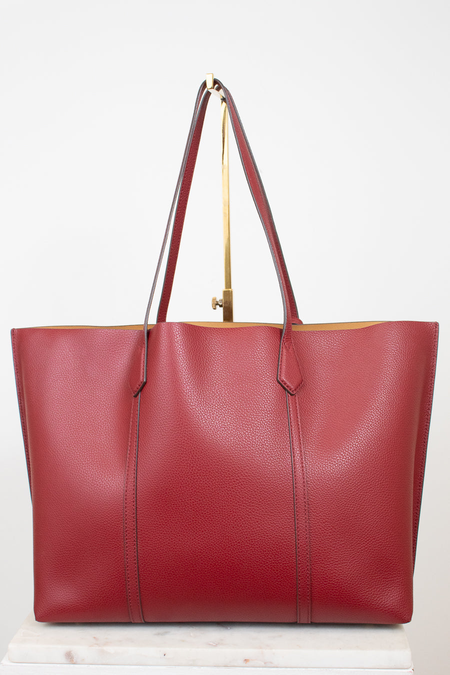 A red pebbled leather tote bag with dark red handles and gold-tone hardware, displayed on a white marble block against a white background.