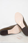 A pair of dark brown leather loafers shown from the bottom, resting on a white marble surface against a white background. The light beige insoles and dark brown outsoles are visible, with a gold-colored brand stamp on the insole.