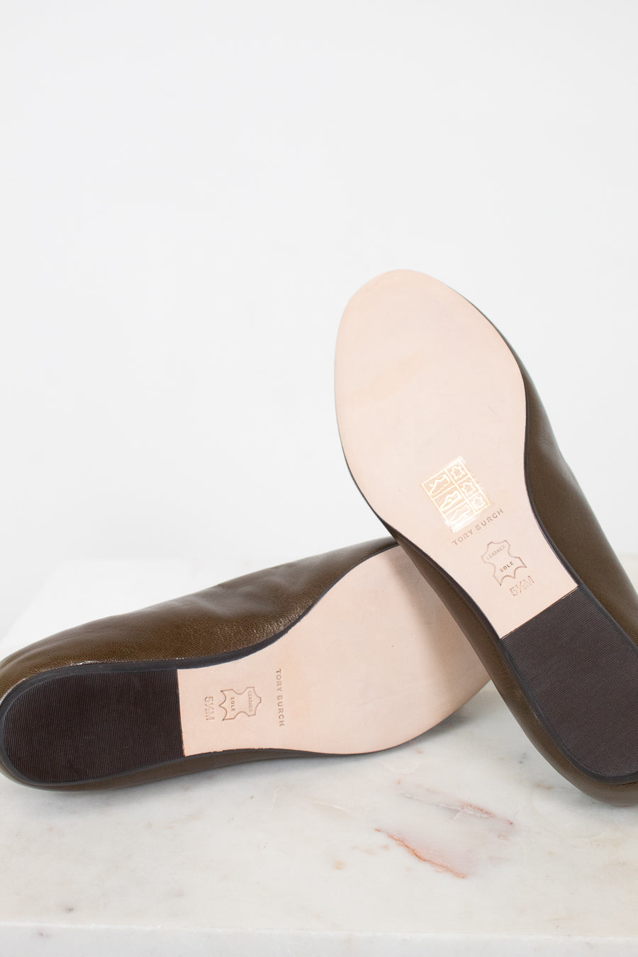 A pair of dark brown leather loafers shown from the bottom, resting on a white marble surface against a white background. The light beige insoles and dark brown outsoles are visible, with a gold-colored brand stamp on the insole.
