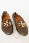 A pair of brown leather Tory Burch loafers with signature gold-tone metal logo hardware, displayed on a white marble surface.