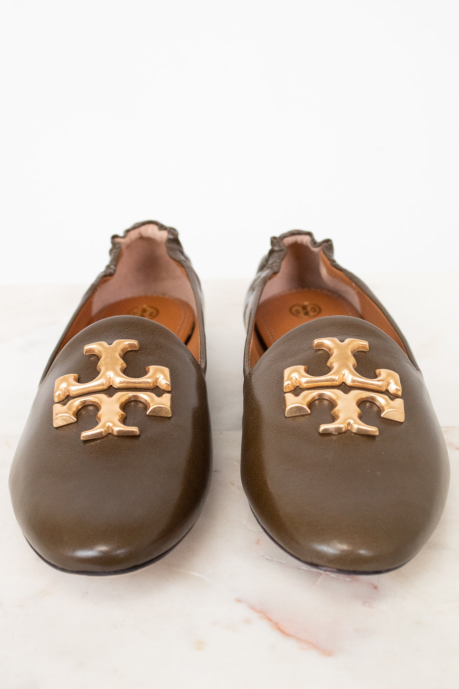 A pair of dark brown leather flats featuring the signature gold Tory Burch logo on the toe, set against a clean white background.