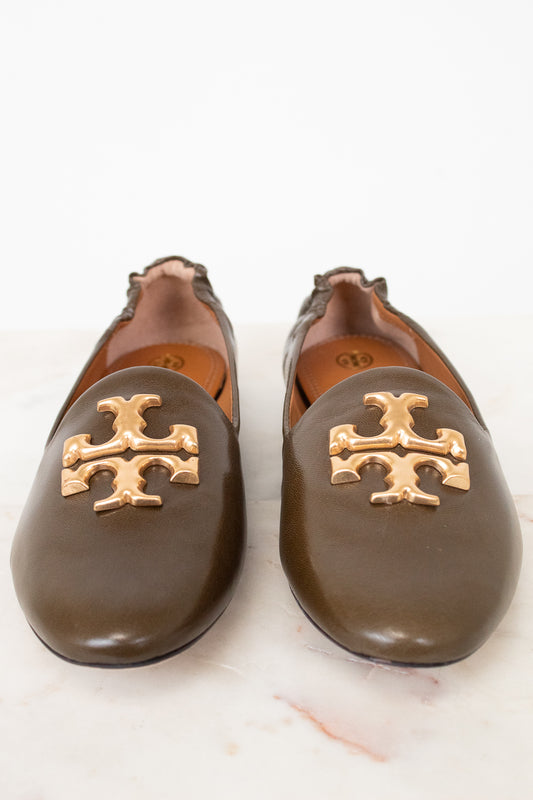 A pair of dark brown leather flats featuring the signature gold Tory Burch logo on the toe, set against a clean white background.