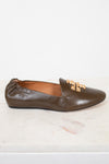 A single brown leather loafer flat with a gold-tone metal logo detail on the toe, displayed on a white marble surface against a white background.