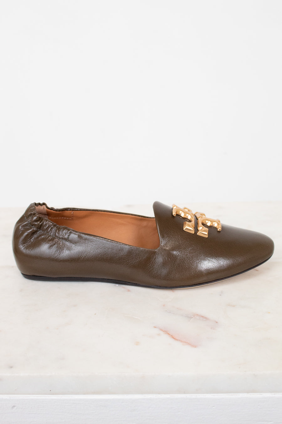 A single brown leather loafer flat with a gold-tone metal logo detail on the toe, displayed on a white marble surface against a white background.