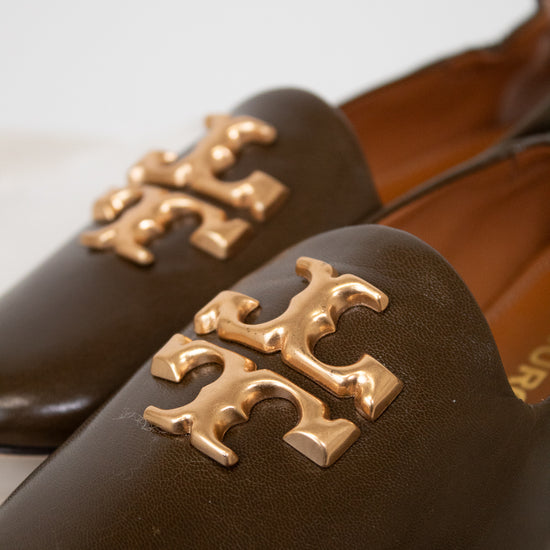 A close-up of a pair of dark brown leather Mary Jane flats featuring a prominent gold-tone metallic Tory Burch logo on the toe box, set against a light neutral background.