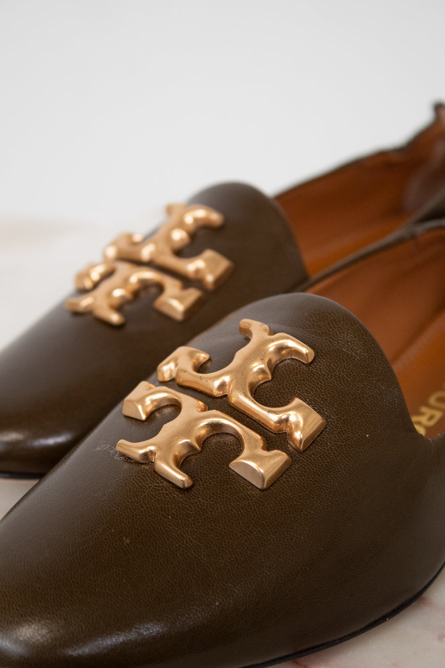 A close-up of a pair of dark brown leather Mary Jane flats featuring a prominent gold-tone metallic Tory Burch logo on the toe box, set against a light neutral background.