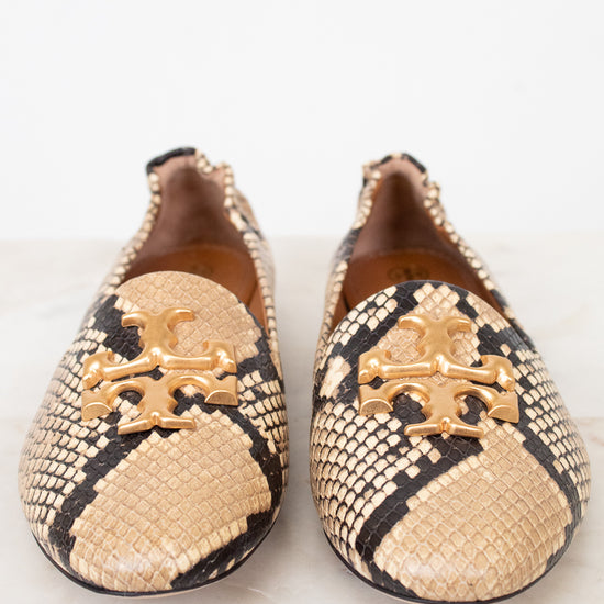 A pair of Tory Burch snakeskin loafers with gold-tone metal logo hardware, displayed on a light marble surface against a white background.
