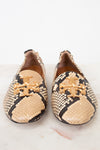 A pair of Tory Burch snakeskin loafers with gold-tone metal logo hardware, displayed on a light marble surface against a white background.