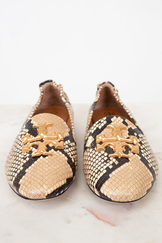 A pair of Tory Burch snakeskin loafers with gold-tone metal logo hardware, displayed on a light marble surface against a white background.