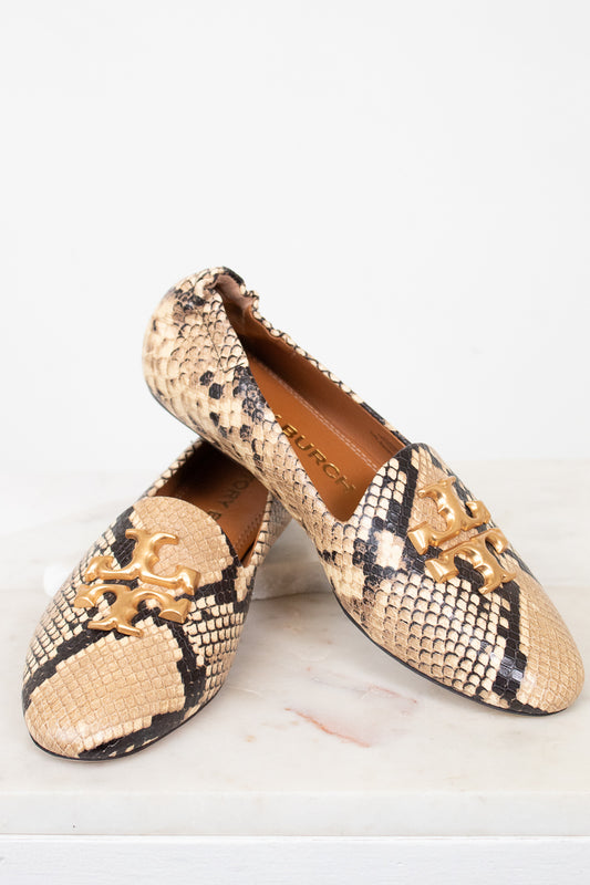 A pair of Tory Burch snakeskin loafers with gold-tone metal logo hardware, displayed on a light marble surface against a white background.