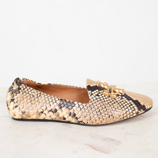 A single women's flat shoe with a beige and black snakeskin pattern and a gold-tone chain detail on the toe, shown on a white marble surface against a white background.