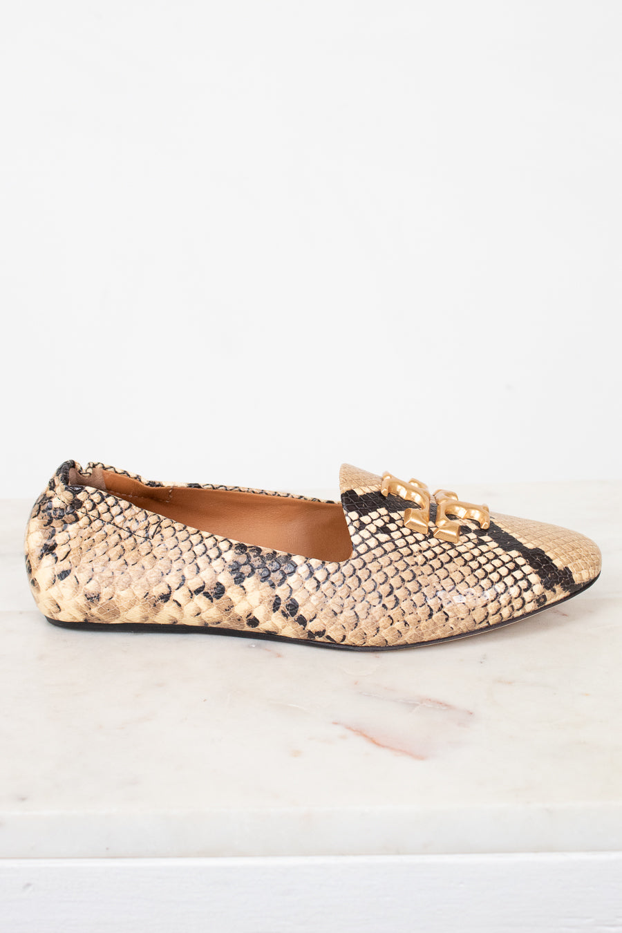 A single women's flat shoe with a beige and black snakeskin pattern and a gold-tone chain detail on the toe, shown on a white marble surface against a white background.