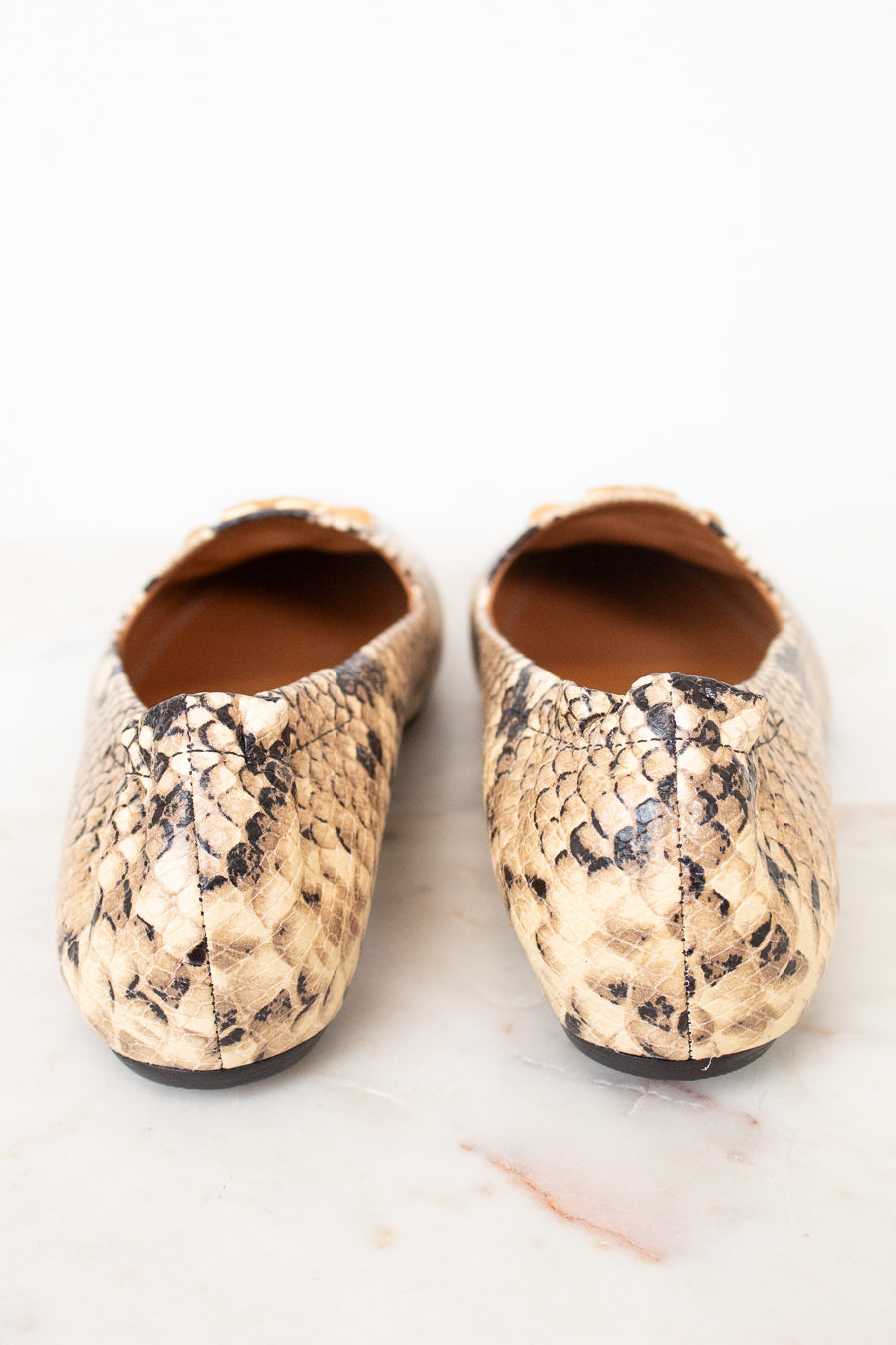 A pair of women's ballet flats featuring a beige and black python skin pattern, photographed from a top-down angle on a light marble surface.