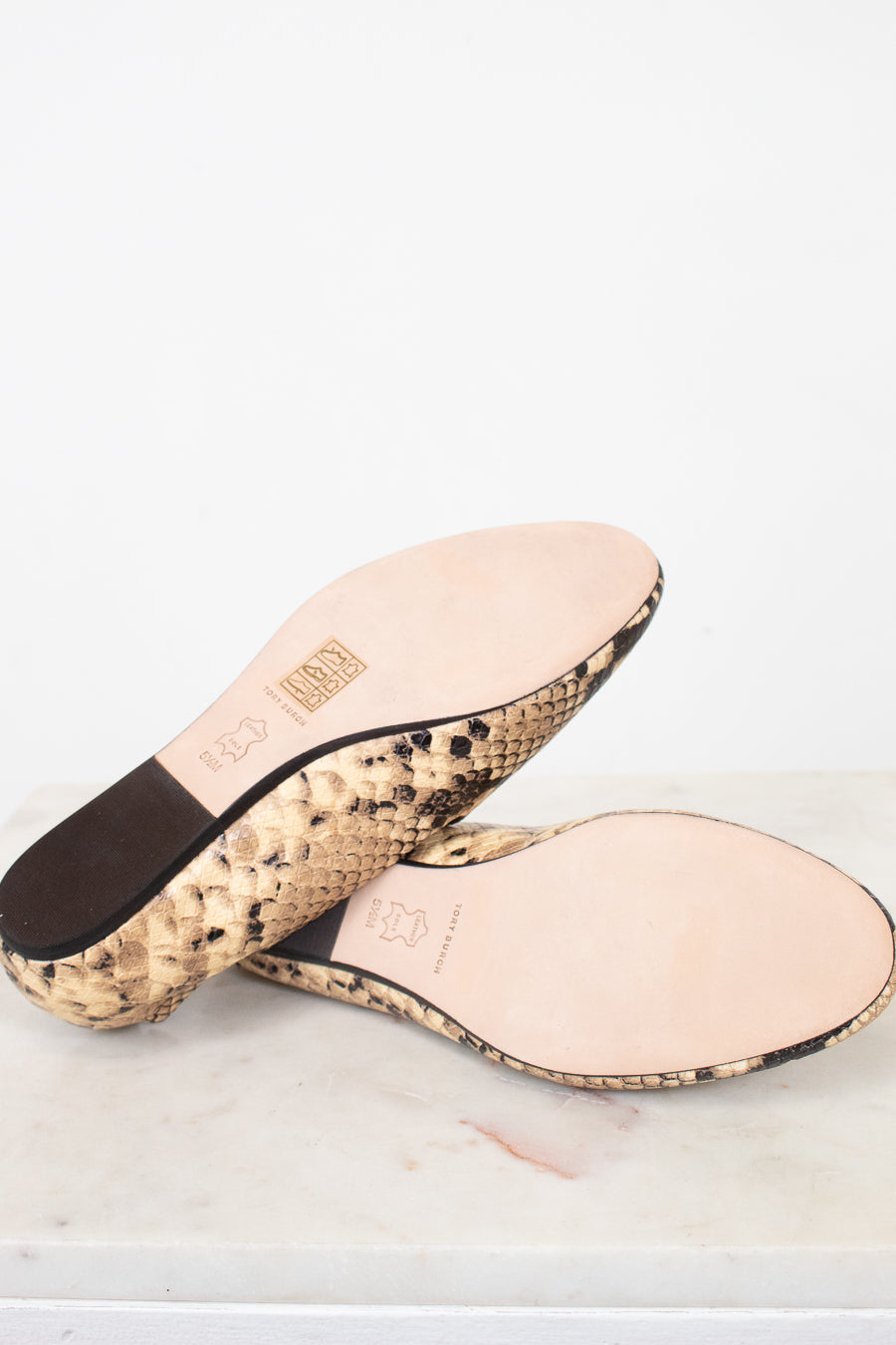 A pair of women's slide sandals shown from the bottom, featuring a beige and black snakeskin patterned sole and a light pink leather insole with embossed branding.