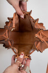 Brown leather handbag with intricate design held open by a person against a neutral background