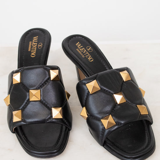 A pair of black leather slide sandals with gold pyramid studs, featuring the Valentino brand name printed on the insole, set against a light marble background.
