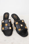 A pair of black leather slide sandals with gold pyramid studs, featuring the Valentino brand name printed on the insole, set against a light marble background.