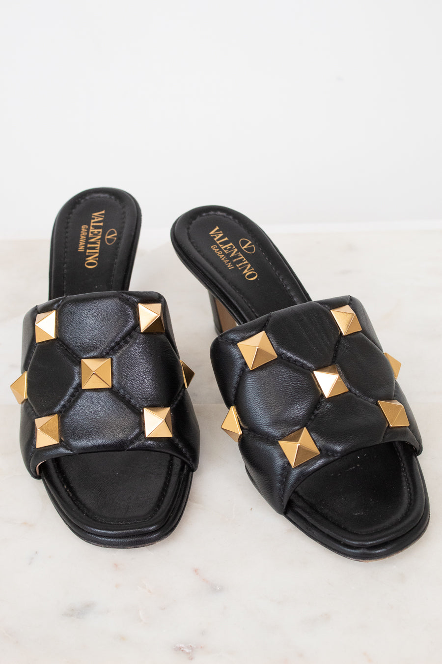 A pair of black leather slide sandals with gold pyramid studs, featuring the Valentino brand name printed on the insole, set against a light marble background.