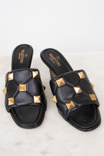 A pair of black leather slide sandals with gold pyramid studs, featuring the Valentino brand name printed on the insole, set against a light marble background.