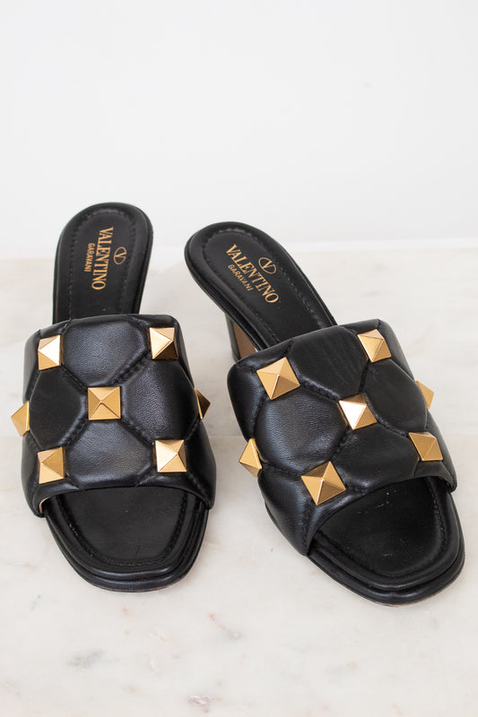 A pair of black leather slide sandals with gold pyramid studs, featuring the Valentino brand name printed on the insole, set against a light marble background.