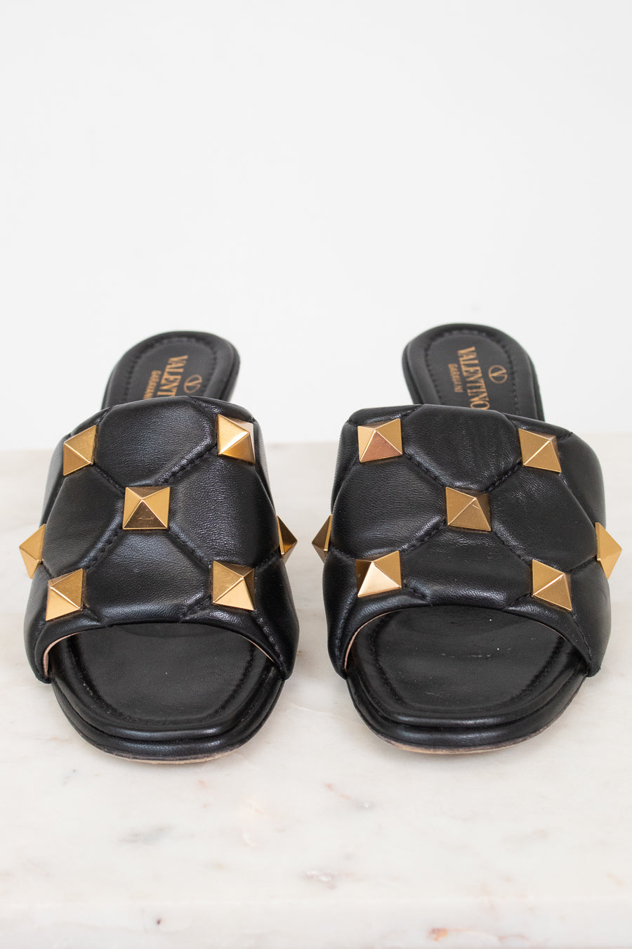 A pair of black leather slide sandals featuring gold pyramid studs and a quilted pattern, with the brand name Valentino visible on the insole.