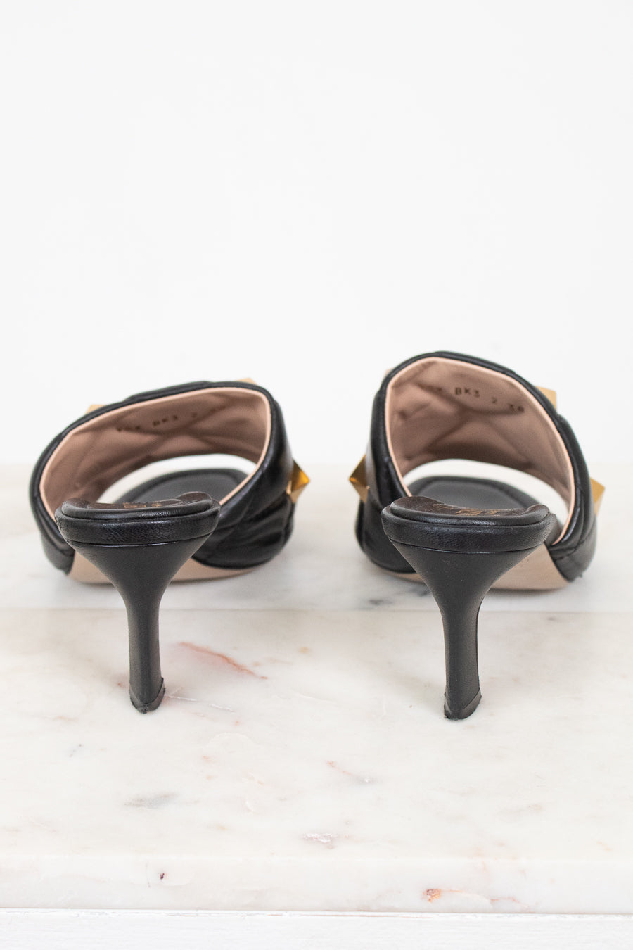 A pair of black leather slingback kitten heels with gold-tone hardware, shown from the back on a white marble surface against a white background.