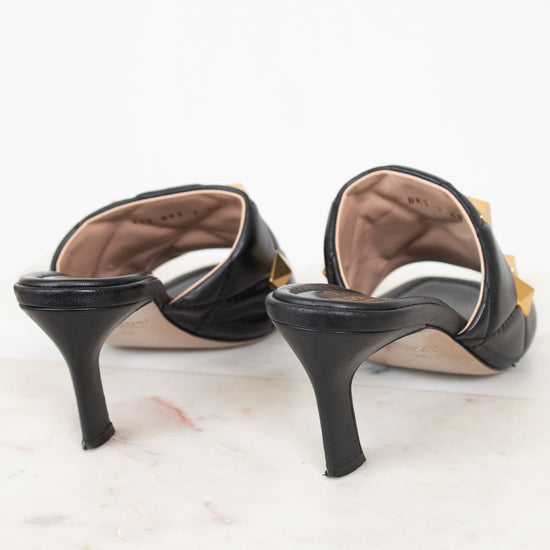 A pair of black leather high-heeled mule sandals with a pointed toe and a gold-tone decorative accent on the outer side, shown from a rear three-quarter angle on a white marble surface against a white background.