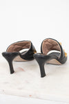 A pair of black leather high-heeled mule sandals with a pointed toe and a gold-tone decorative accent on the outer side, shown from a rear three-quarter angle on a white marble surface against a white background.
