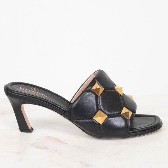 A single black leather mule sandal with a low block heel and gold pyramid stud embellishments, shown on a white marble surface against a white background.