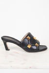 A single black leather mule sandal with a low block heel and gold pyramid stud embellishments, shown on a white marble surface against a white background.