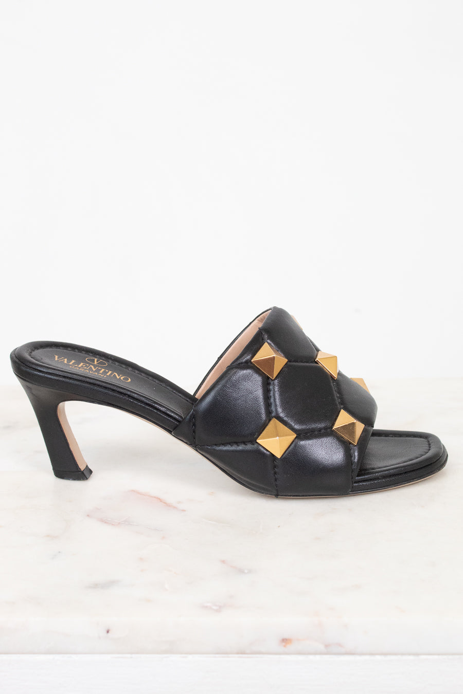 A single black leather mule sandal with a low block heel and gold pyramid stud embellishments, shown on a white marble surface against a white background.