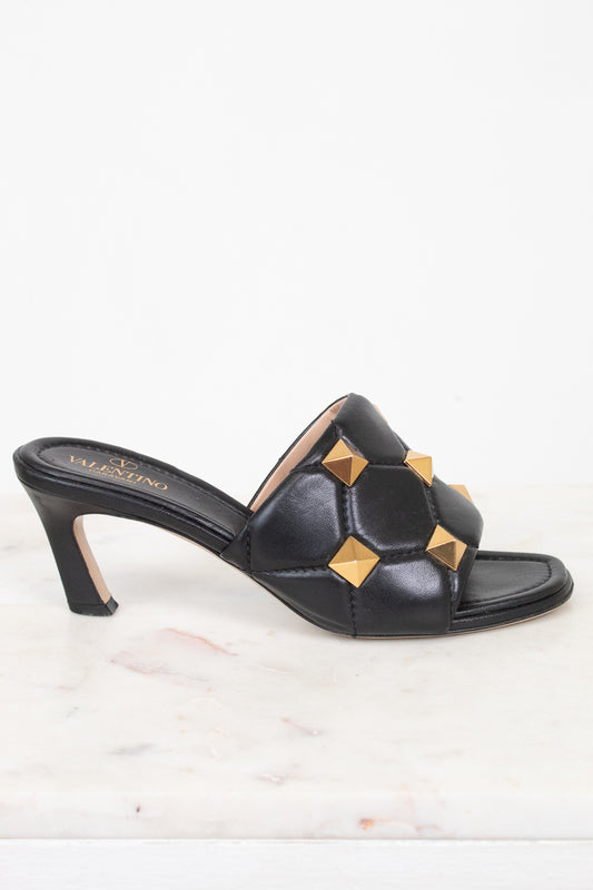 A single black leather mule sandal with a low block heel and gold pyramid stud embellishments, shown on a white marble surface against a white background.