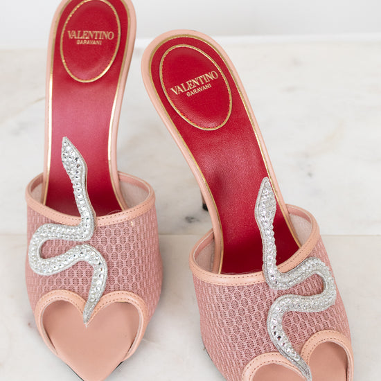 Pink high-heeled shoes with rhinestone snake detail on a white background with Valentino logo.