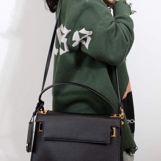 Person wearing a green sweatshirt with white text, holding a black handbag against a plain background