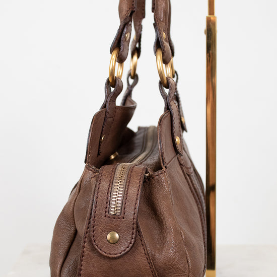 Brown leather handbag with handles on a gold stand against a white background