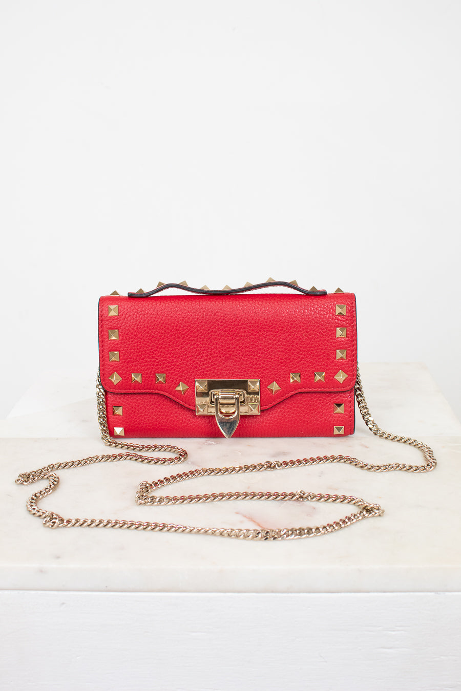 Red rectangular leather bag against a plain  background. Bag is decorated with gold-tone rock-stud details and a chain as well as a studded top handle.