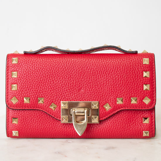 Close-up of red rectangular leather bag decorated with gold-tone rock-stud details and top handle against a plain background