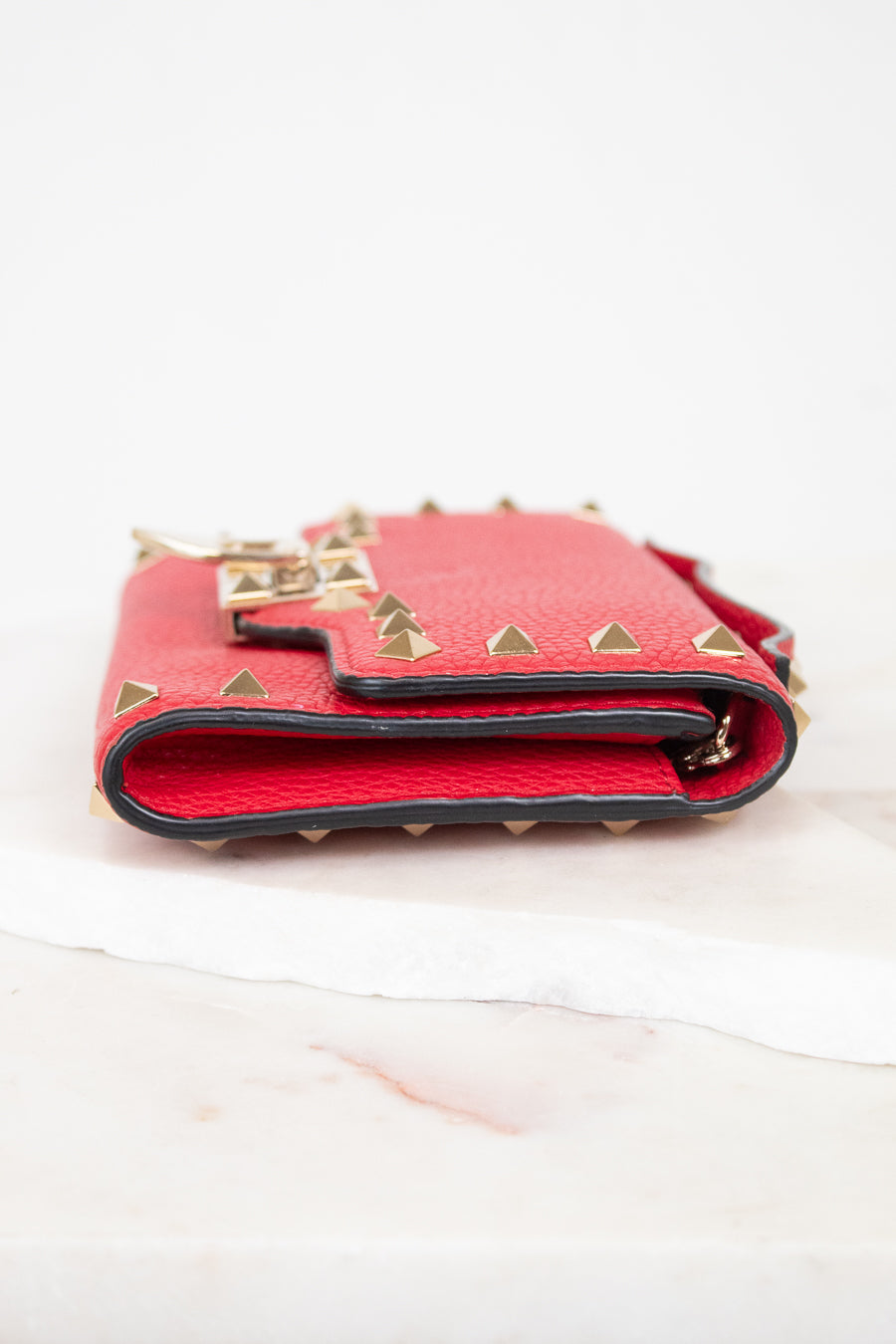 Side view of red rectangular leather bag decorated with gold-tone rock-stud details laying on marble surface.