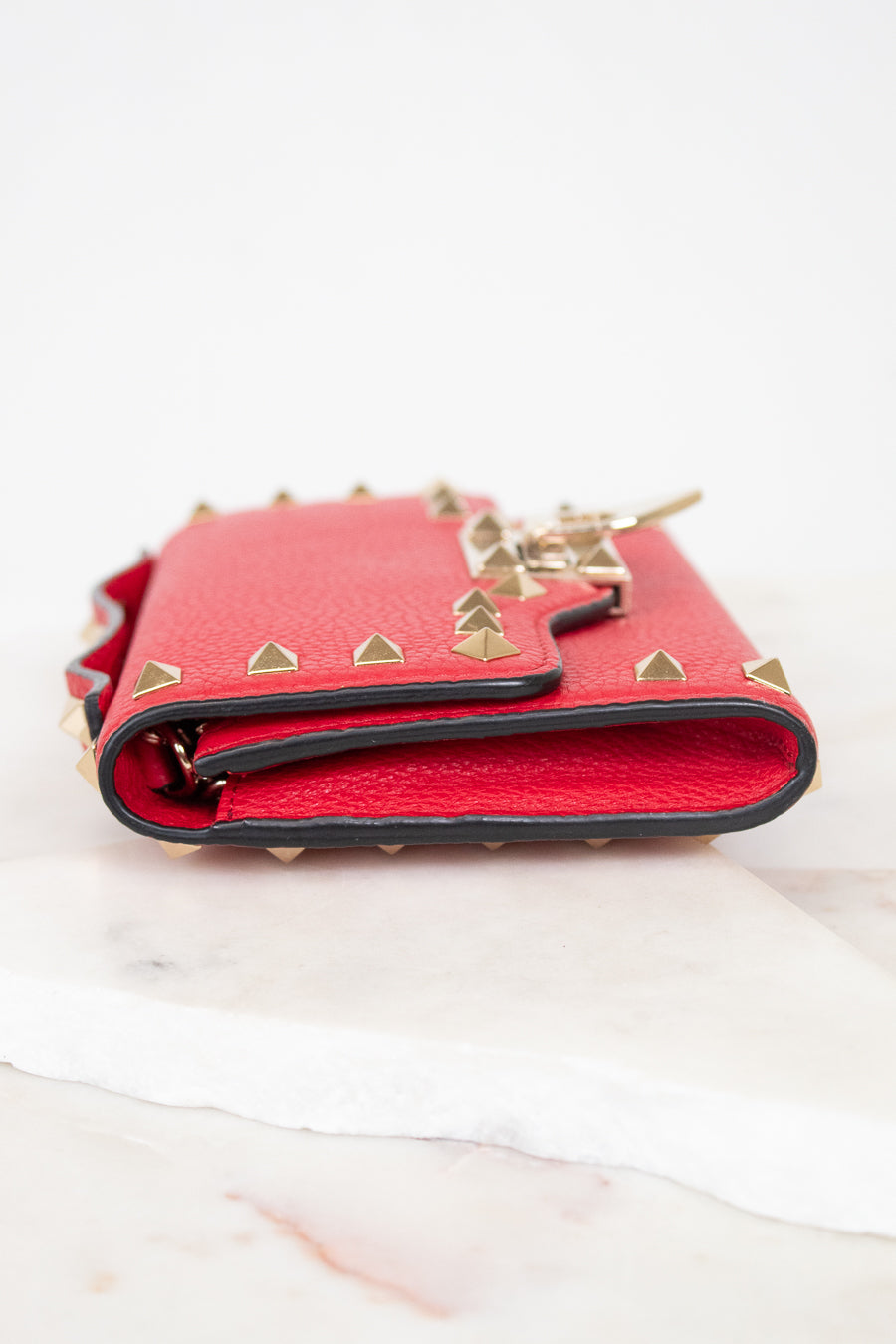 Side view of red rectangular leather bag decorated with gold-tone rock-stud details laying on marble surface.