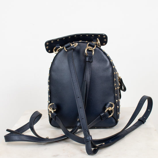 Back view of a navy blue leather mini backpack with gold-tone hardware and studded details, on a white marble surface against a white background.