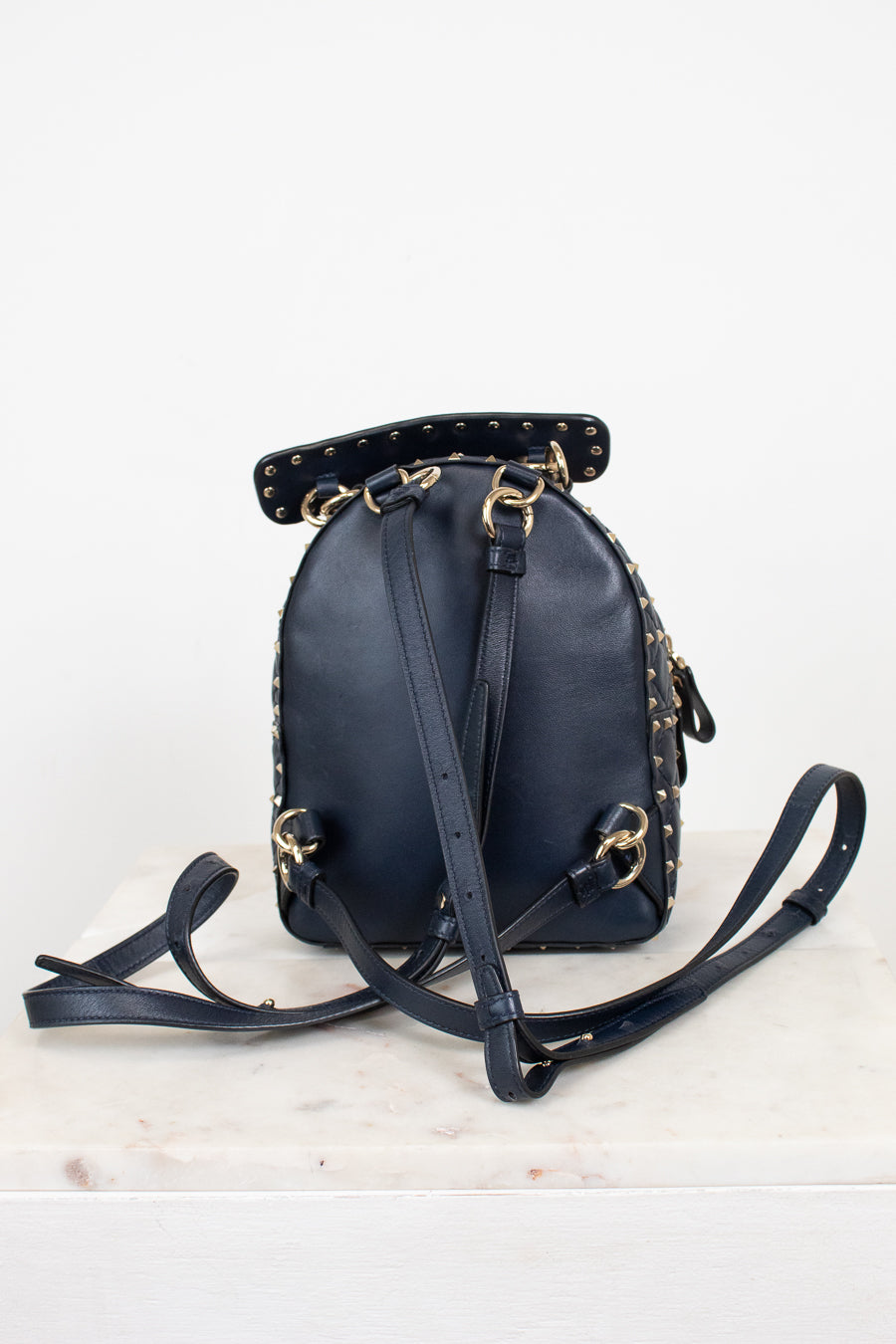 Back view of a navy blue leather mini backpack with gold-tone hardware and studded details, on a white marble surface against a white background.