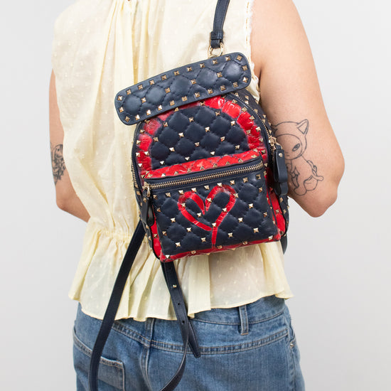 A person shown from the back wearing a small quilted backpack with navy and red panels, metal studs, and a red heart motif on the front, paired with a cream sleeveless top and blue jeans.