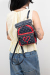 A person shown from the back wearing a small quilted backpack with navy and red panels, metal studs, and a red heart motif on the front, paired with a cream sleeveless top and blue jeans.