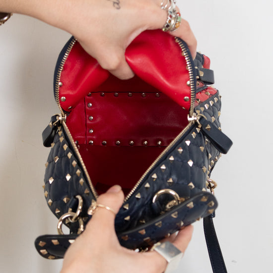 A navy blue studded leather handbag with a bright red interior, shown being opened by two tattooed hands wearing silver rings against a plain white background.