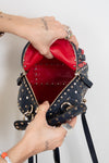 A navy blue studded leather handbag with a bright red interior, shown being opened by two tattooed hands wearing silver rings against a plain white background.