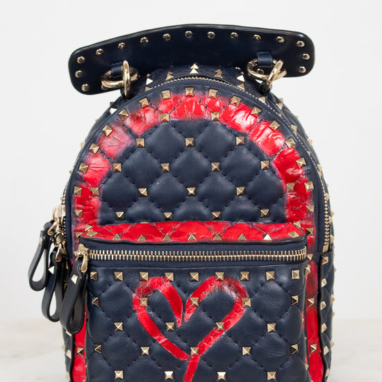 A small quilted backpack with navy blue and red details, gold-tone studs, and a red heart design on the front, photographed against a plain light background.